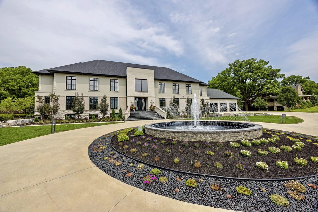 A large two-story house with many windows stands behind a landscaped circular driveway featuring a central fountain and colorful flowers, surrounded by green lawns and trees under a partly cloudy sky.