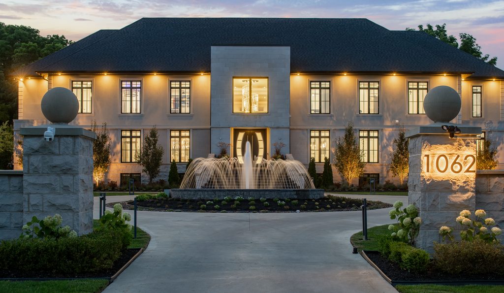 A large, elegant house with many windows is illuminated at dusk. A fountain sits in the center of the driveway, and 1062 is displayed on a stone pillar at the entrance. Lush landscaping surrounds the scene.