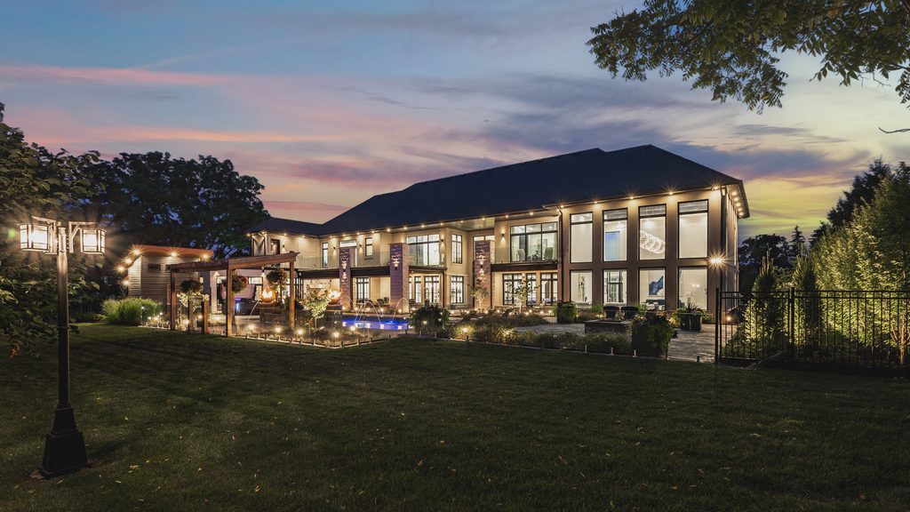 A luxurious modern mansion at twilight, featuring illuminated floor-to-ceiling windows, a swimming pool with fountains, a well-manicured lawn, and a vibrant sunset sky.
