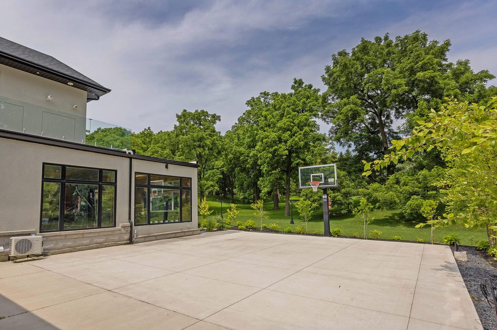A spacious concrete patio beside a modern house with large windows, featuring a basketball hoop in the center. The background shows a lush green lawn surrounded by mature trees under a cloudy sky.