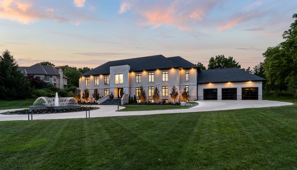Large modern house with illuminated windows, black roof, and attached three-car garage, set on landscaped lawn with a circular driveway and a fountain in front. Trees and sunset sky are visible in the background.