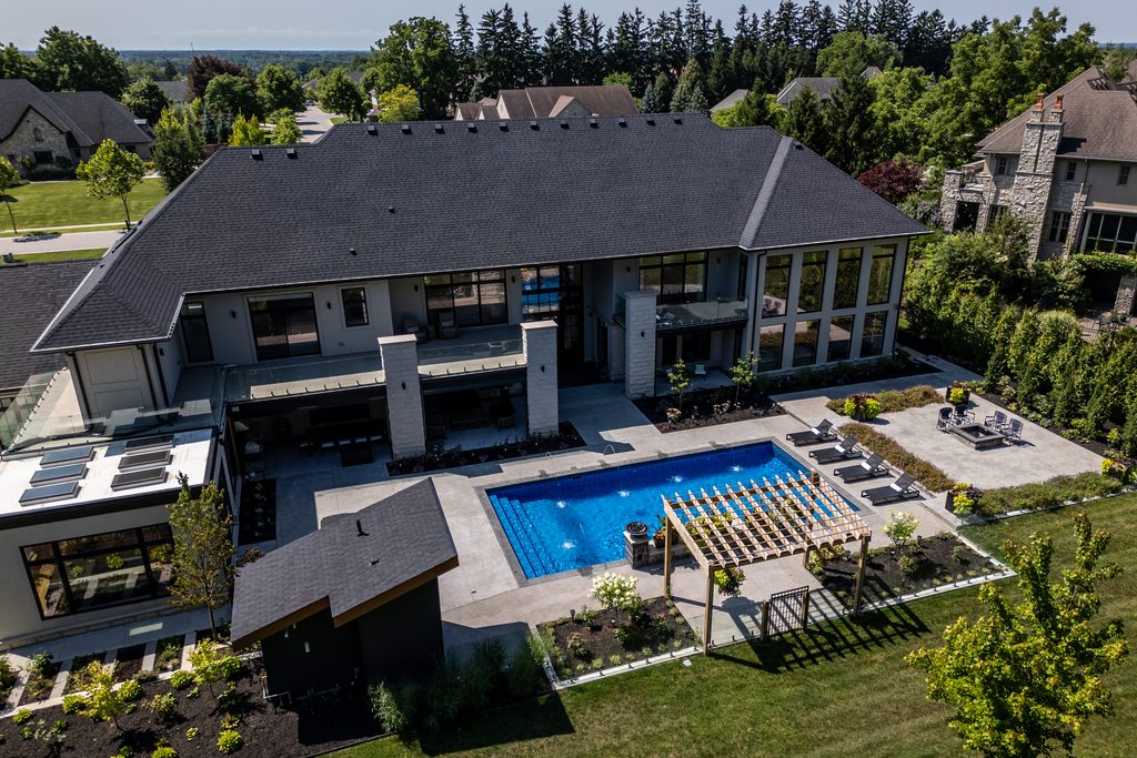 Aerial view of a modern two-story house with large windows, a blue rectangular pool, lounge chairs, a pergola, landscaped yard, and surrounding trees.
