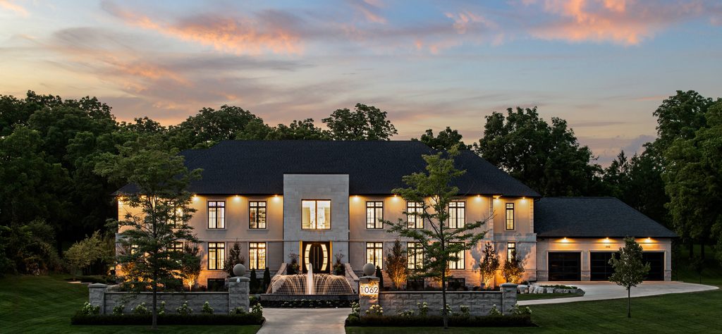 Large, modern mansion with extensive windows, exterior lights, a central fountain, and a three-car garage, surrounded by trees and manicured lawns at sunset.