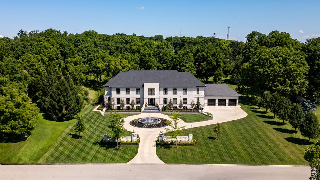 A large, white two-story mansion with a circular driveway, central fountain, manicured lawn, and surrounded by trees under a clear blue sky.