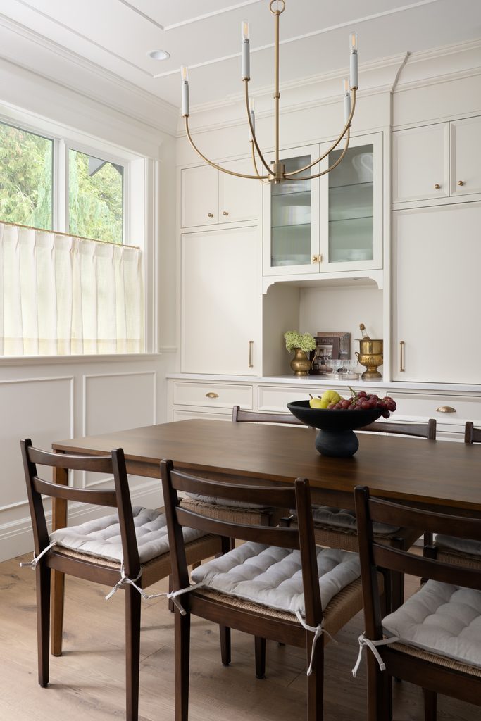 A dining room with a wooden table, six cushioned chairs, a black fruit bowl, white cabinetry, gold accents, a modern chandelier, and a window with sunlight filtering through light curtains.