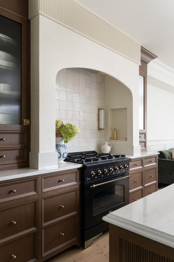 A modern kitchen with wood cabinets, a black stove, white tile backsplash, marble countertops, and decorative vases, all under a large arched hood.