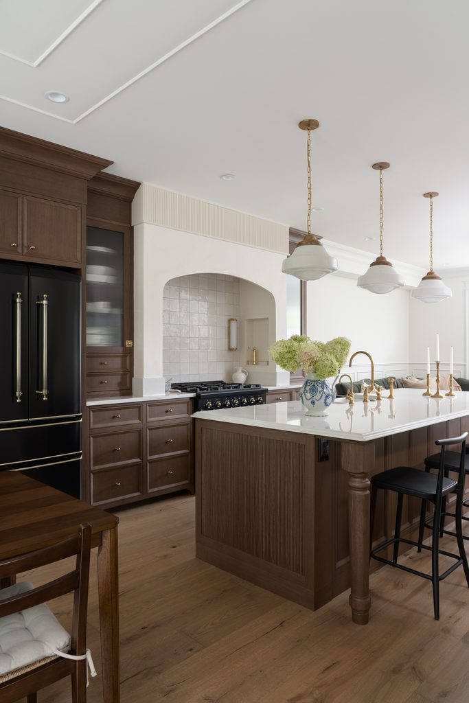 A modern kitchen with wood cabinets, a black refrigerator, a white island with a sink, gold fixtures, pendant lights, and a vase of green flowers. There are black chairs and a wooden dining table in the foreground.