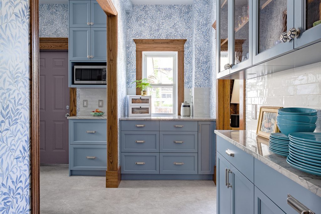 A bright kitchen with blue cabinets, wood trim, marble countertops, blue patterned wallpaper, a small window, and teal dishes on display.
