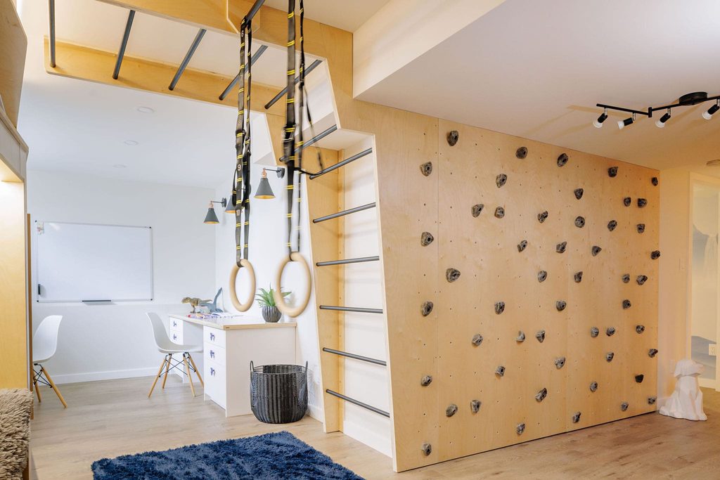 A modern room with a climbing wall, gymnastics rings, and a small desk area. Light wood tones, white accents, and a blue rug create a bright, playful atmosphere for work and indoor activity.