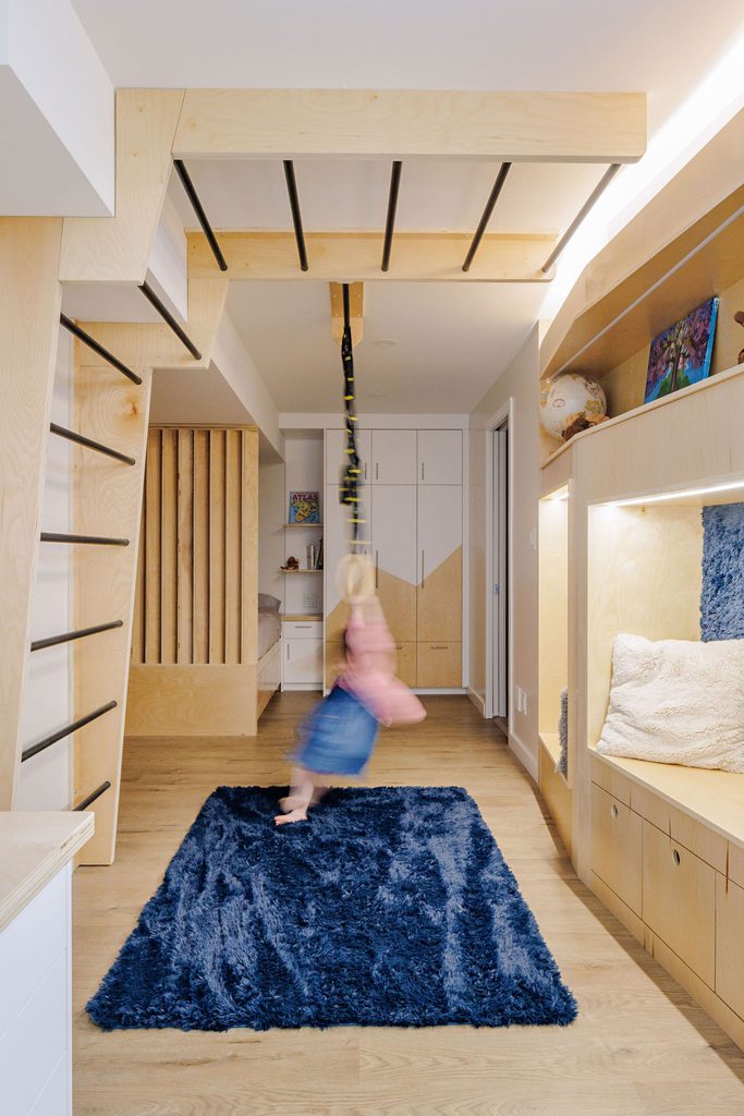 A child swings from monkey bars in a modern, bright room with wood accents, built-in storage, and a blue rug.
