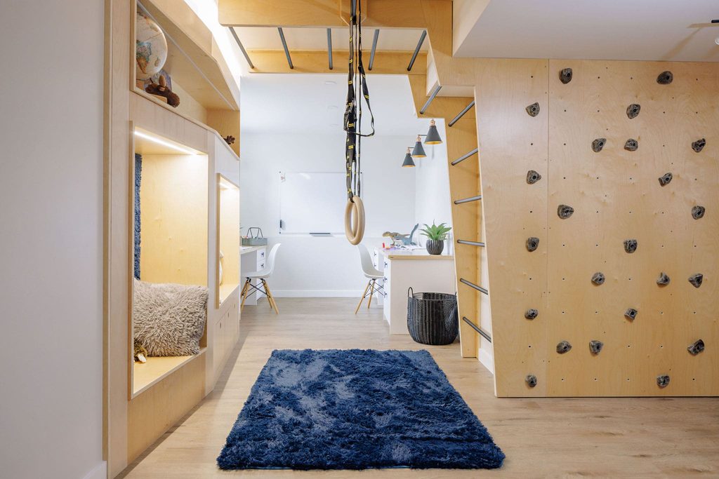 A modern playroom with a climbing wall, ladder, gymnastic rings, cozy built-in nook with pillows, blue rug, wooden floor, and a desk area in the background.