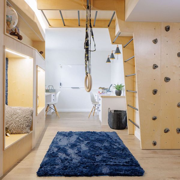 A modern playroom with a climbing wall, ladder, gymnastic rings, cozy built-in nook with pillows, blue rug, wooden floor, and a desk area in the background.