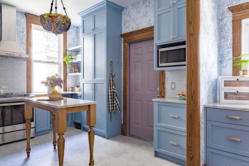 A colorful kitchen with blue cabinets, a central wooden island, marble countertops, and blue-and-white leaf-patterned wallpaper. A mauve door, stained wood trim, a stained-glass pendant light, and stainless steel appliances complete the scene.