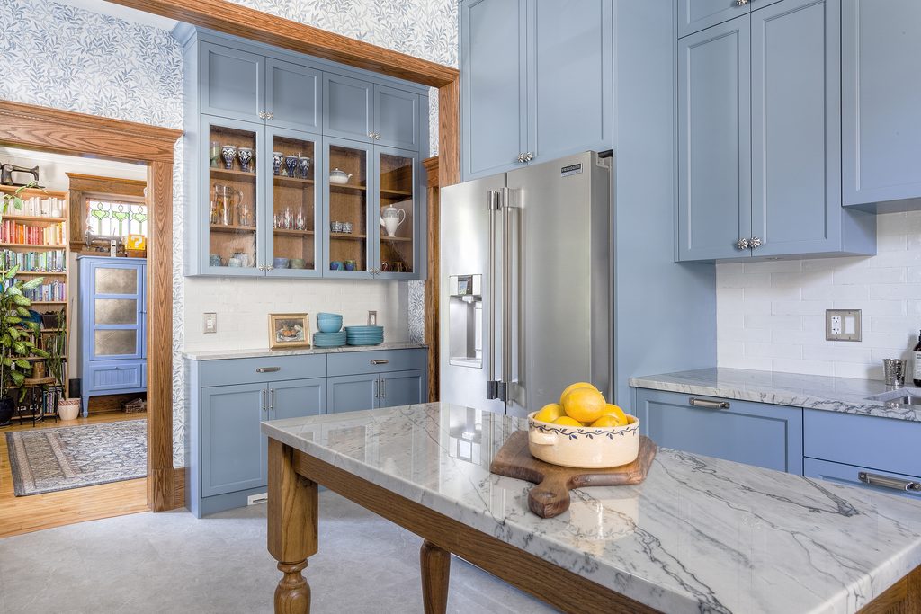 Bright kitchen with light blue cabinets, stainless steel fridge, marble island countertop, and a wooden bowl of lemons. Glass-front cabinets display dishes. View into another room with bookshelves and plants.
