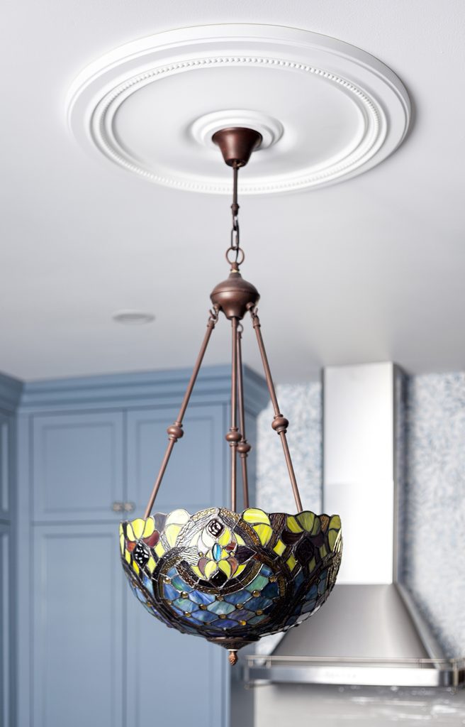 A stained glass pendant light with a bronze fixture hangs from a white ceiling medallion in a kitchen with blue cabinets and patterned wallpaper.