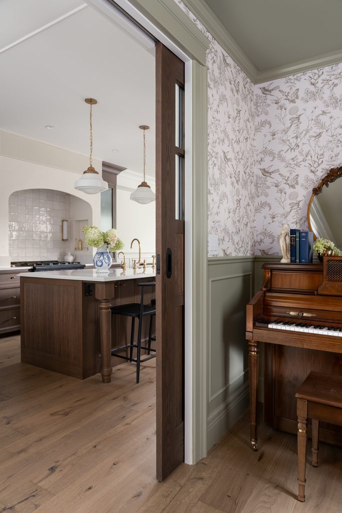 A cozy kitchen with wood accents is visible through a sliding door; in the foreground, a wooden piano with books and a vase of flowers sits against a floral-patterned wall.