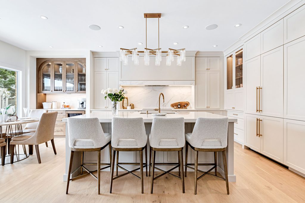 Modern kitchen with white cabinets, a marble backsplash, a central island with four upholstered barstools, gold fixtures, pendant lighting, and a dining nook by a large window.