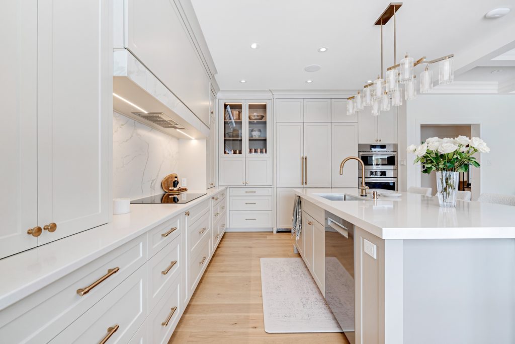 Modern white kitchen with gold accents, marble backsplash, built-in appliances, an island with a sink, a flower vase, and pendant lighting. Light wood flooring and minimal, clean design throughout.