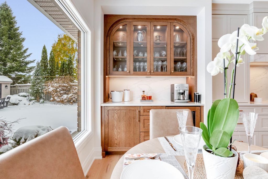 A bright kitchen with a wooden cabinet displaying glassware, a coffee machine on the counter, and a dining table set for a meal. A large window shows a snowy backyard with trees and patio furniture.