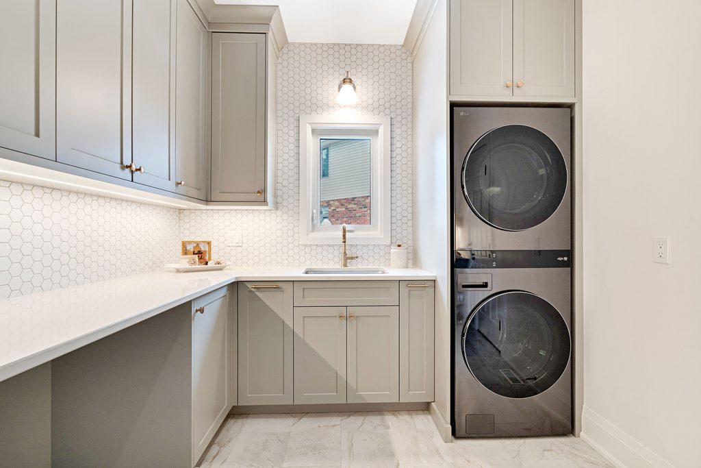 Modern laundry room with gray cabinets, white hexagon tile backsplash, stacked black washer and dryer, a window above the sink, and bright lighting.