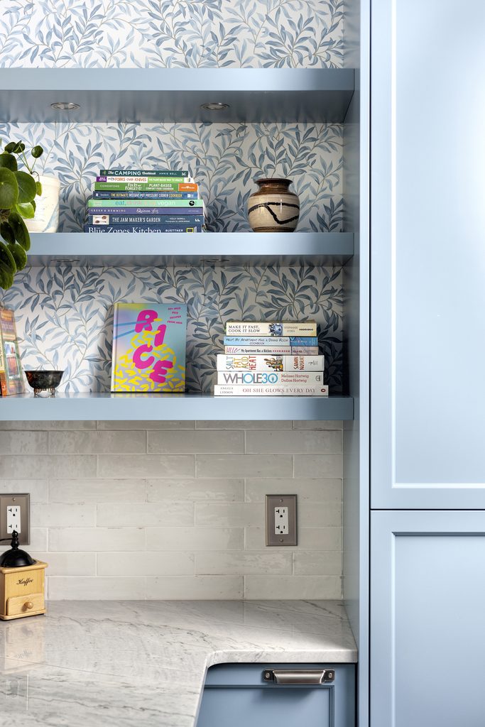 Open blue shelves with cookbooks, a plant, and a mug, set against a blue leaf-patterned wallpaper above a white tile backsplash and light marble countertop in a kitchen.