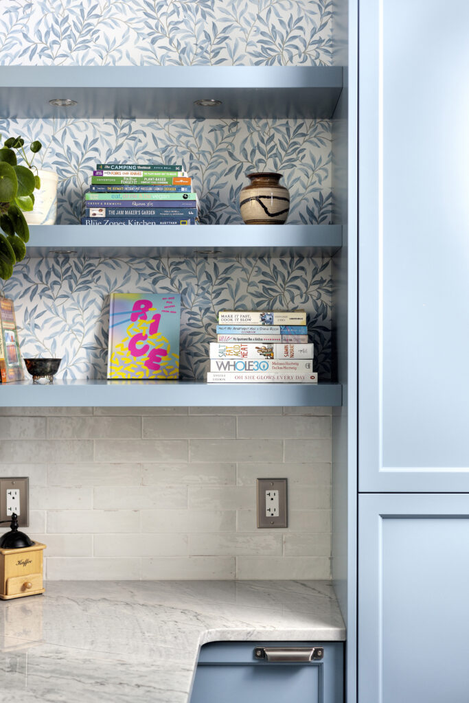 Blue kitchen shelves holding books, a plant, and a small ceramic pot. The wall has blue leaf-patterned wallpaper and white tile. Light blue cabinets and a gray marble countertop are also visible.