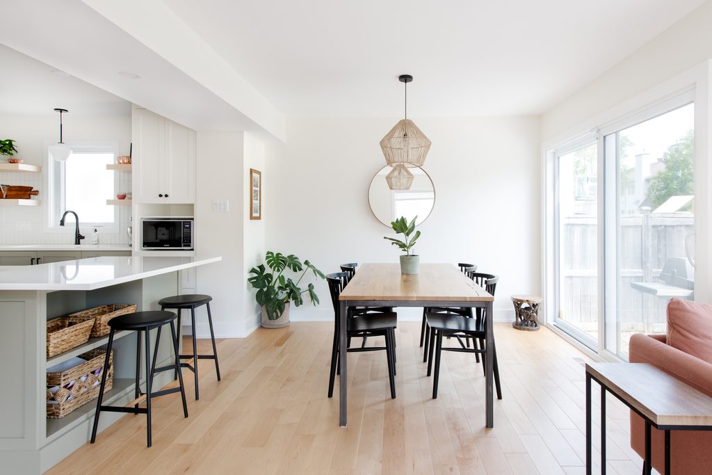 Modern dining area with a wooden table, black chairs, a round mirror, hanging wicker lamps, and plants. Large glass doors let in natural light; kitchen with bar stools is visible on the left.