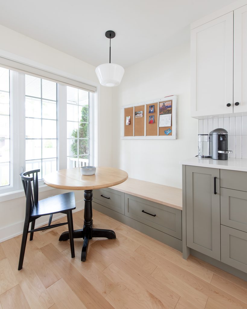 A bright kitchen nook with a round wooden table, black chair, built-in bench, coffee maker, and a corkboard on the wall. Large windows let in natural light.