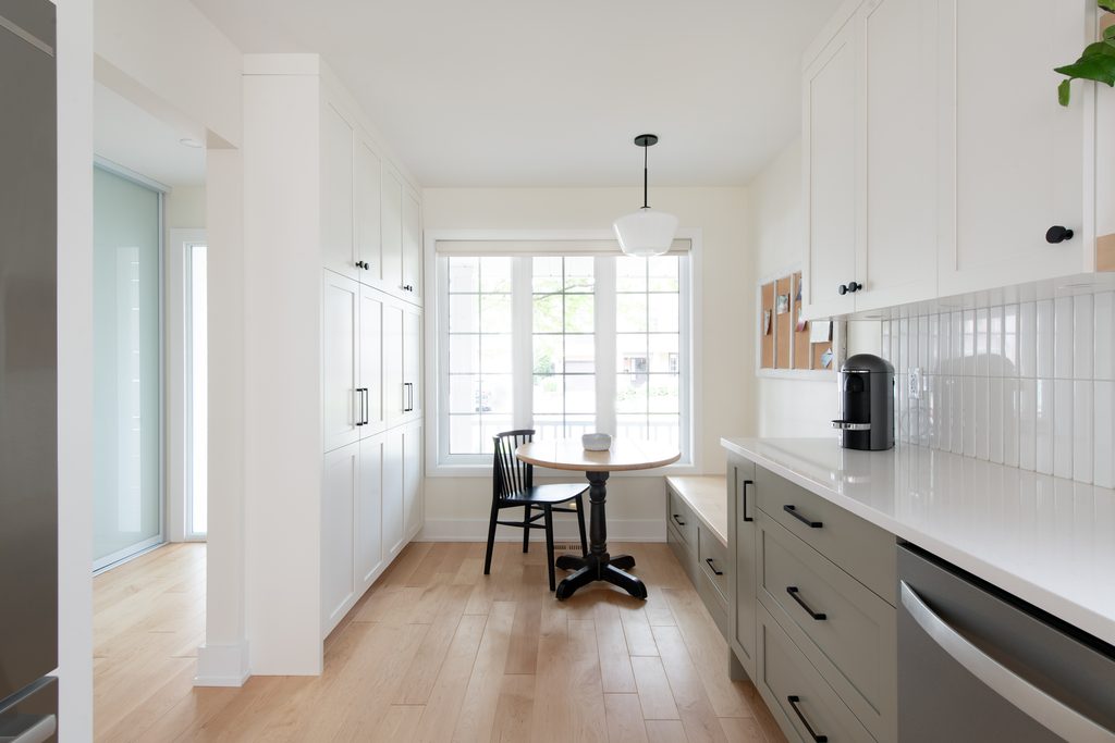 Bright kitchen nook with white cabinets, light wood floor, a small round table and two chairs by a large window. A coffee maker sits on the counter, and cork boards hang on the wall.