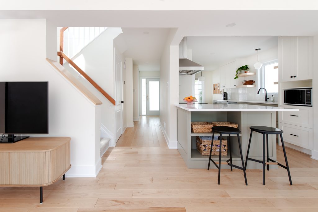Bright modern kitchen with a central island, two black stools, white cabinets, and open shelves. Light wood floors and stairs lead to a hallway and glass door. A TV sits on a wooden stand to the left.