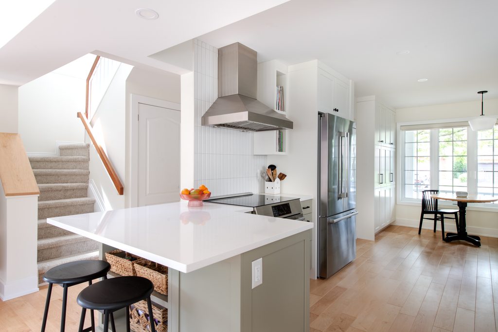 Bright, modern kitchen with white cabinets, stainless steel appliances, a center island with black stools, and a bowl of fruit. Stairs and a dining area with large windows are visible. Light wood flooring throughout.