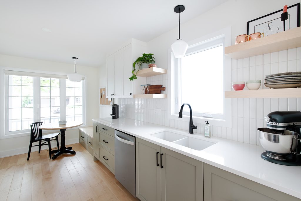 A bright, modern kitchen features light sage green lower cabinets, white upper cabinets, white quartz countertops, and a white tile backsplash. A dining nook with a small round table and black chair sits by a large window.