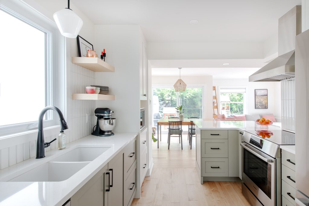 Bright modern kitchen with white cabinets, light wood floor, stainless steel appliances, open shelves, and a window over a double sink. Dining area with large windows is visible in the background.