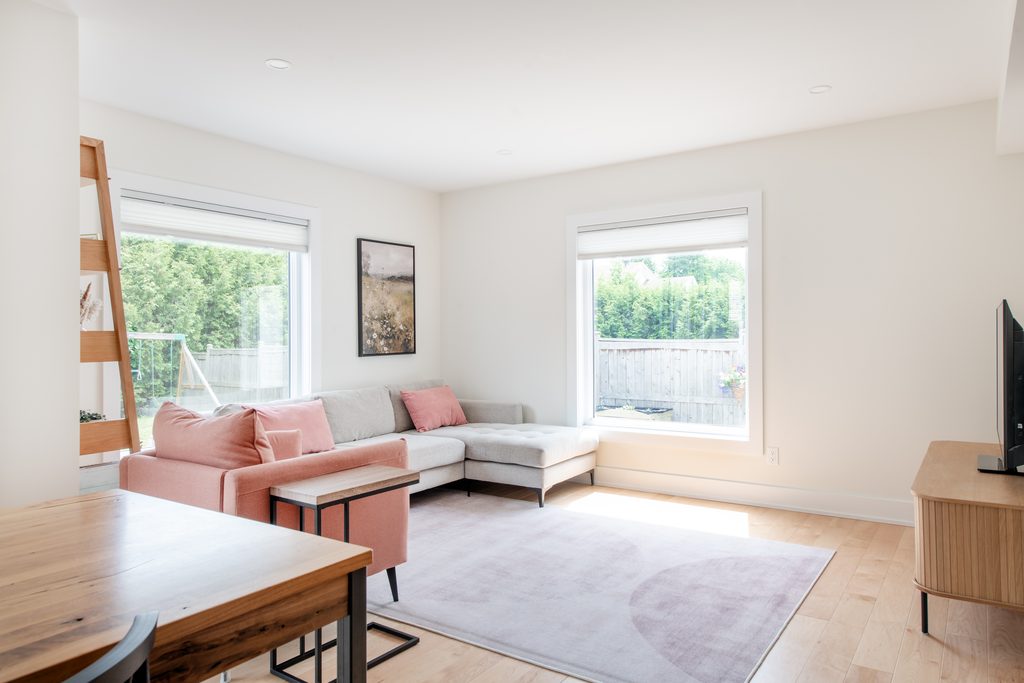 Bright living room with a gray sectional sofa, pink cushions, a light purple rug, a wooden table, and large windows letting in sunlight. A TV sits on a wooden stand across from the sofa.