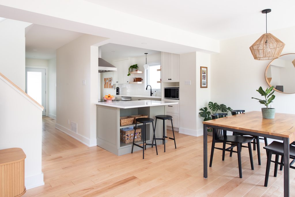 Bright, modern kitchen and dining area with light wood floors, white cabinets, a small kitchen island with stools, dining table with black chairs, plants, and a round mirror on the wall.