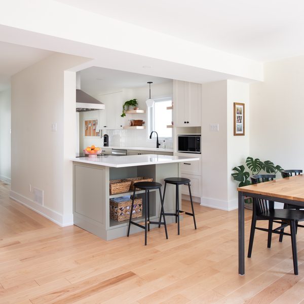 Bright, modern kitchen and dining area with light wood floors, white cabinets, a small kitchen island with stools, dining table with black chairs, plants, and a round mirror on the wall.