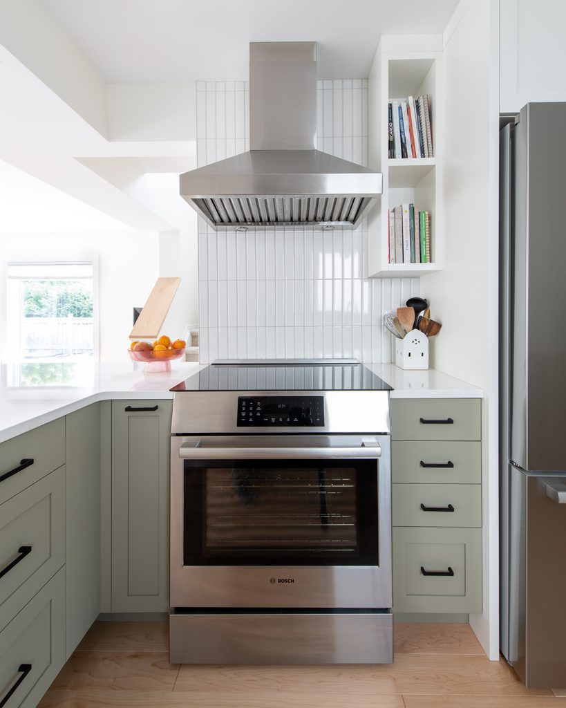 Modern kitchen with stainless steel oven and range hood, light green cabinets, white tile backsplash, and open shelves with books and utensils. Bowl of fruit sits on the counter by a large window.