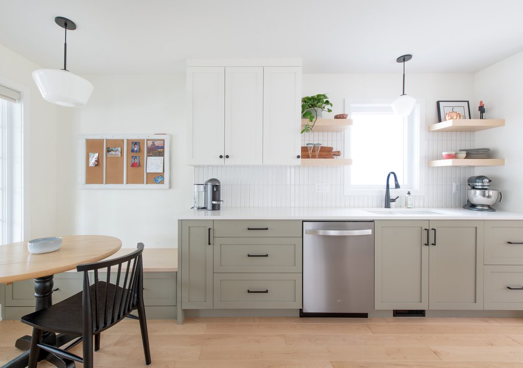Bright, modern kitchen with white cabinets, light green lower drawers, stainless steel dishwasher, open shelves, plants, and a small round dining table with two chairs near a window.