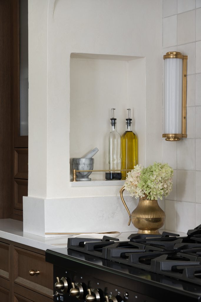 A kitchen nook with olive oil and vinegar bottles, a marble mortar and pestle, a brass pitcher with white hydrangeas, and a gas stovetop in the foreground.