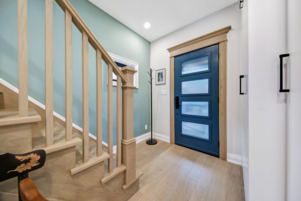 A modern entryway with light wood flooring, a blue front door with glass panels, mint green accent wall, wooden staircase, and a black coat rack.