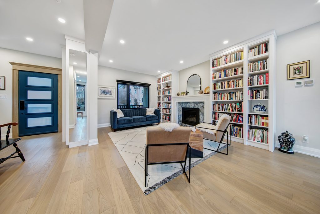 Modern living room with light wood floors, a navy couch, two tan chairs, a fireplace, built-in bookshelves, and a blue front door. Large windows and recessed lighting create a bright, open atmosphere.