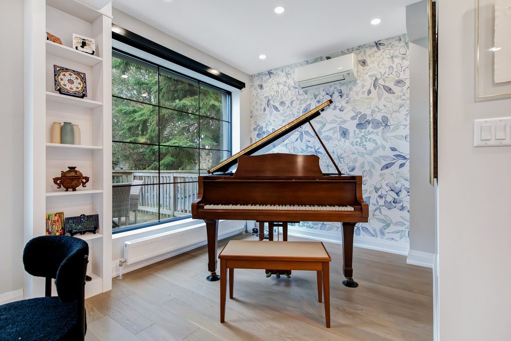 A grand piano with a bench sits by a large window overlooking trees, in a bright room with floral wallpaper, wood floors, and built-in shelves displaying decor items.