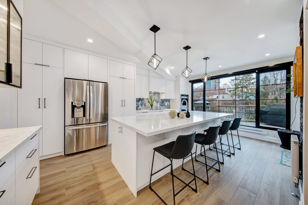Modern kitchen with white cabinets, stainless steel appliances, island with marble countertop, four black barstools, pendant lights, wood flooring, and large windows with outdoor view.