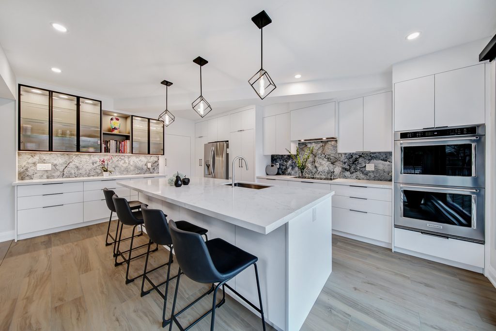 Modern kitchen with white cabinets, marble countertops, black bar stools, stainless steel appliances, and pendant lights. Wood flooring and a marble backsplash add elegance. Open shelving displays decor items.
