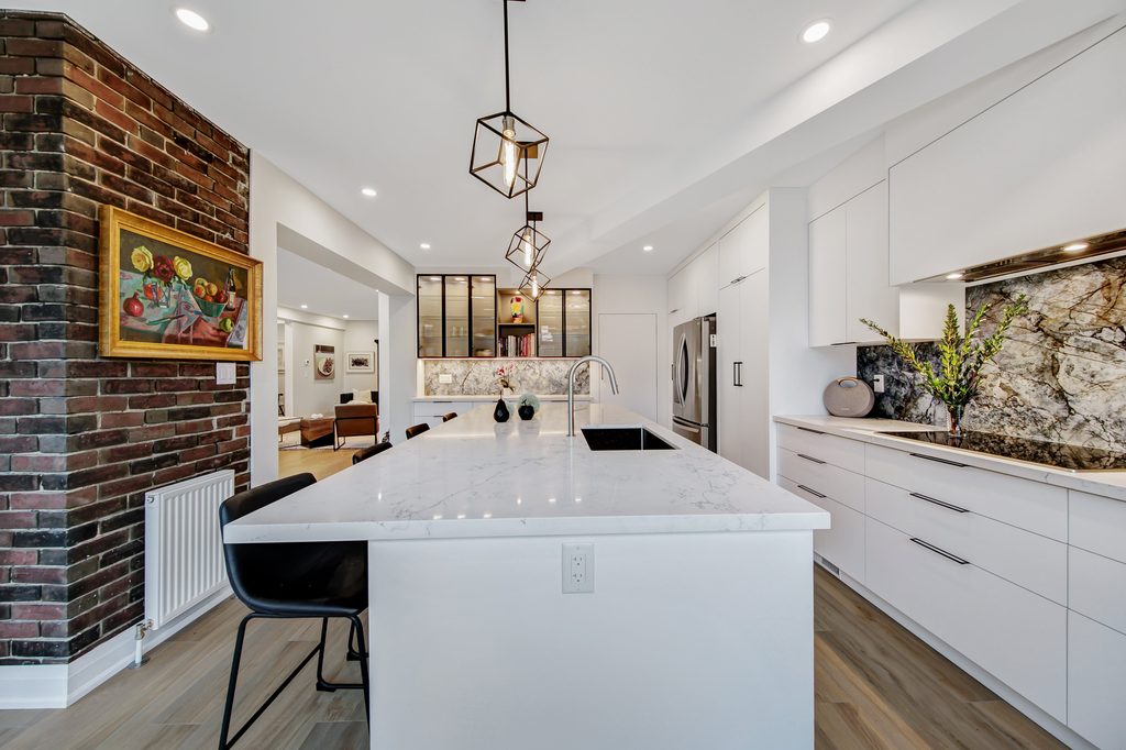 Modern kitchen with white cabinets, marble countertops, geometric pendant lights, a brick accent wall with art, and a vase with flowers on the counter. Dining area and living space are visible in the background.