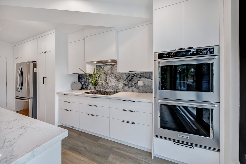 Modern kitchen with white cabinets, stainless steel double wall ovens, a marble countertop and backsplash, built-in refrigerator, and wood flooring. A vase with greenery is placed on the counter near the cooktop.