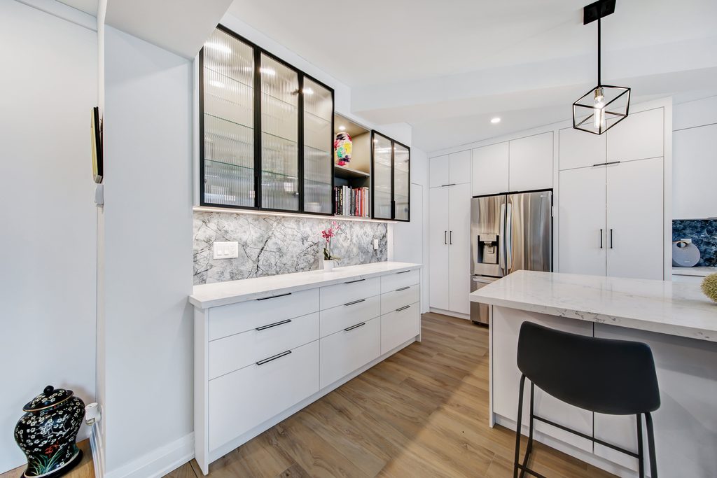 Modern kitchen with white cabinets, marble backsplash, glass-front upper cabinets, stainless steel refrigerator, island with a white countertop, black chair, and wood flooring. Decorative vase and pink flowers add color.
