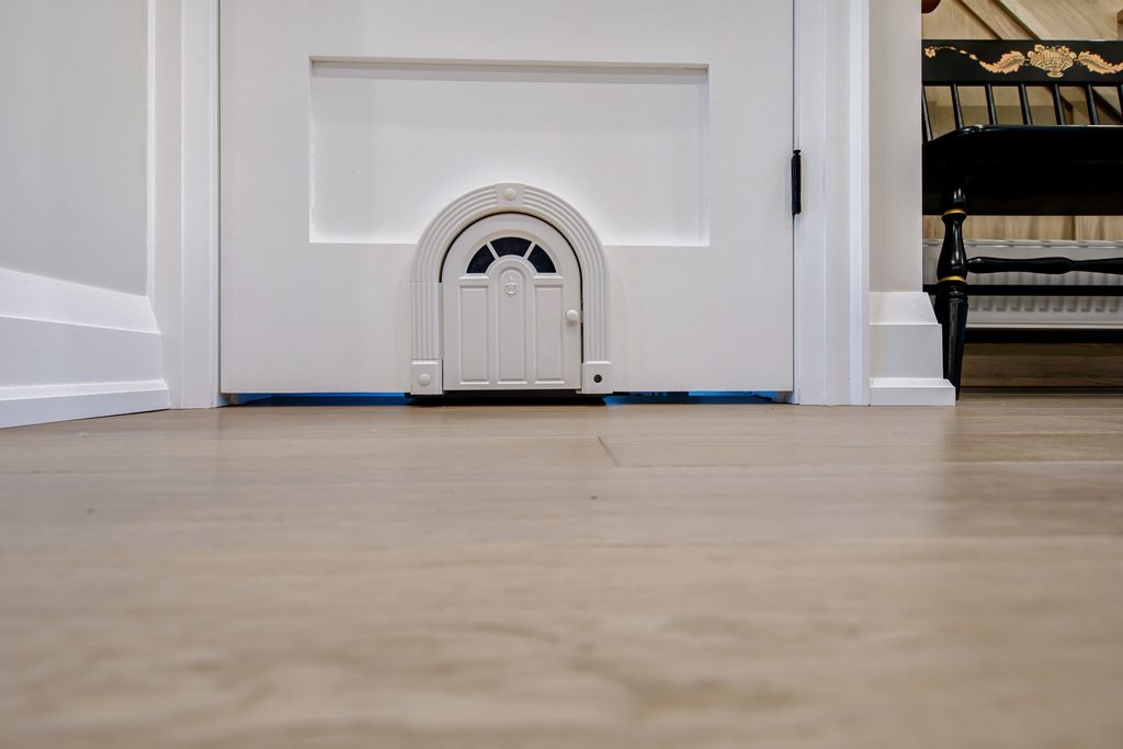 A small, decorative door is installed at the base of a larger white door in a bright room with wood floors. A black chair with gold accents is partially visible in the background.