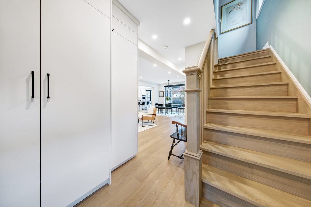 A modern hallway with light wood stairs, white cabinets on the left, and a view into an open living area with large windows, chairs, and a dining table in the background.