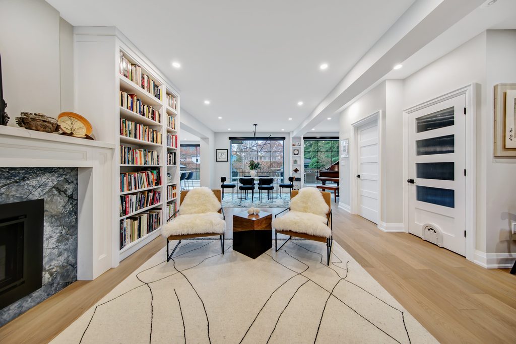 Modern living room with two chairs, a bookshelf, a fireplace, and a dining area in the background. Large windows let in natural light, and wood floors add warmth to the space.